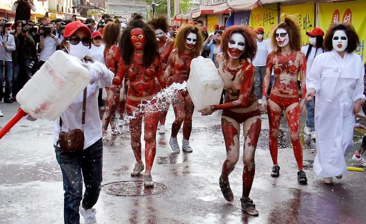 Eight women covered in red body paint and white face paint, with two of them in wearing white clothing, pour flammable liquid onto the street in front of onlookers.