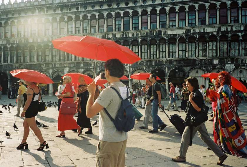 A group of approximately seven people walk past a grand building with arched windows in Venice, holding red umbrellas.