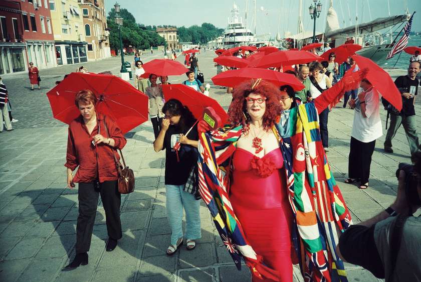 A group of approximately 12 people walk down a sunny street in Venice holding red umbrellas.