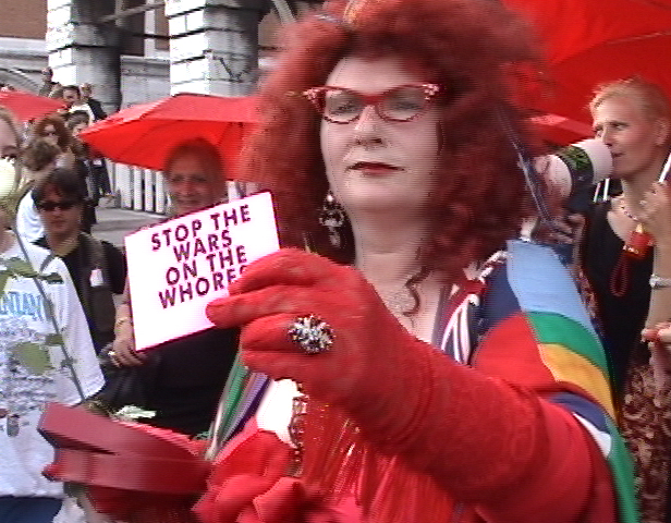 A woman with red hair and glasses is walking down the street. In her red gloved hand she holds a sign that says 'stop the wars on the whores'. People in the background are looking at her.