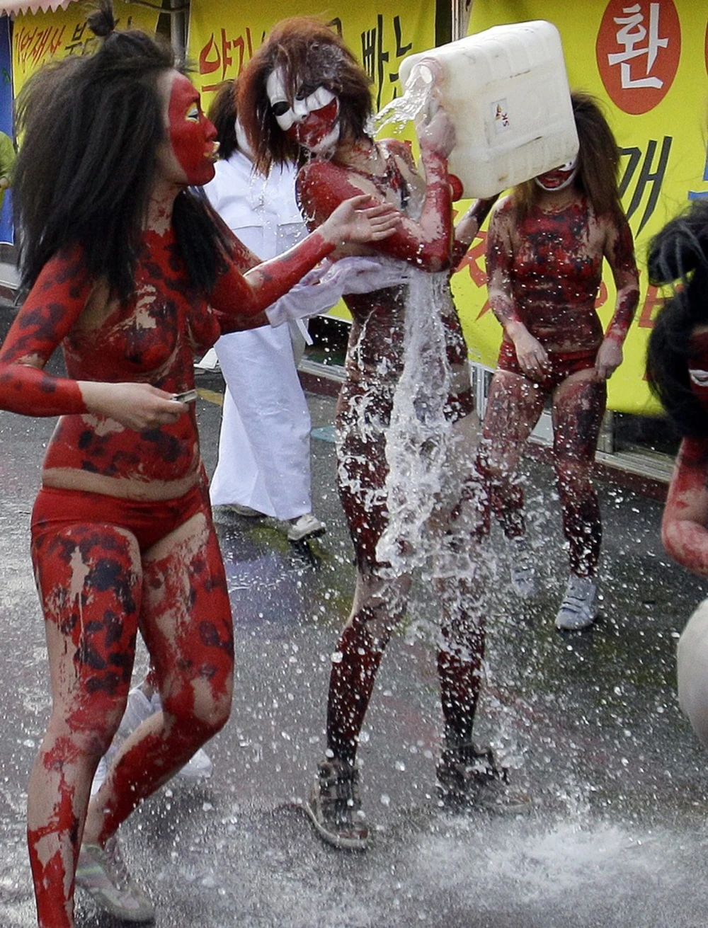 Three women with red body paint, one of them holds a large container of clear liquid and is pouring it onto the ground and onto the person next to them.