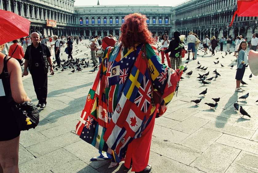 A woman with red hair walks through a plaza on a sunny day in Venice. A large rectangle of fabric is draped over her shoulders that is printed with different flags. Two red umbrellas frame the image on each side.