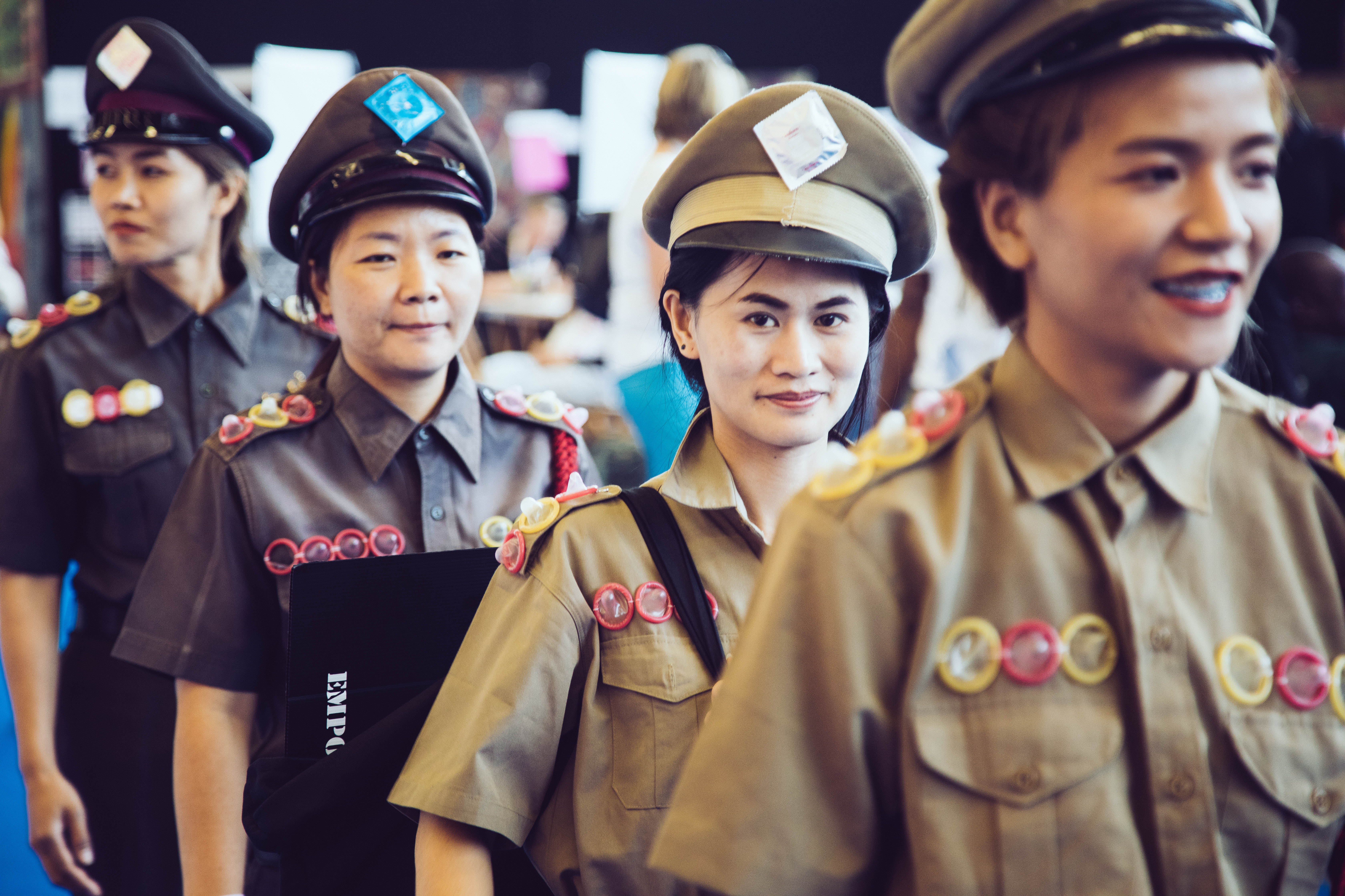 A group of four East Asian woman in military-style outfits decorated with condoms walk through a room.
