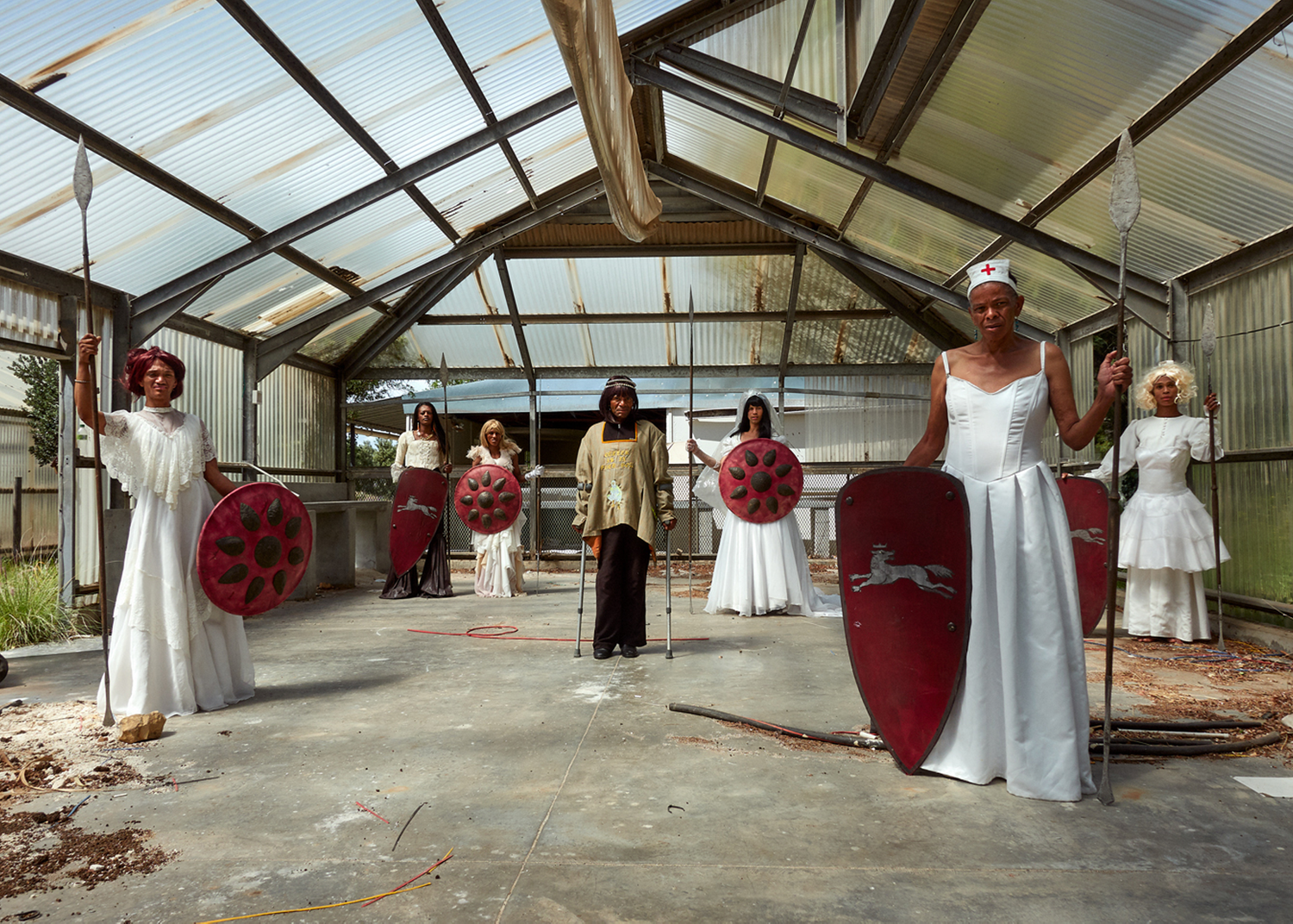 Seven figures are standing in a warehouse with a slanted roof. All but the central figure is wearing white and holding red shields of varying shapes and sizes