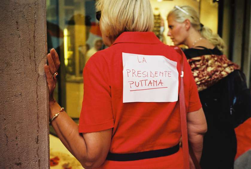 A woman with bobbed blonde hair is turned away from the camera, one hand on the edge of an open door. She's wearing a red top with an a4 sheet of paper on her back that says 'LA PRESIDENTE PUTTANA'
