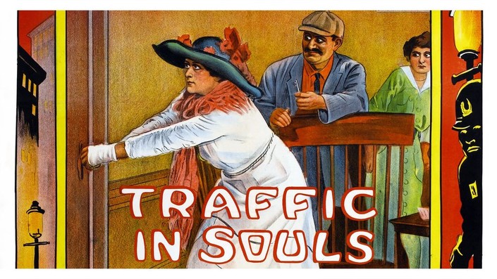 A brightly coloured illustration from 1913 for the film 'Traffic in Souls'. A woman in a white dress and black hat tries to open the door of the room she is trapped inside. Behind her is a man in a blue suit with a moustache and brown skin, as well as another frightened looking white woman in a green dress.