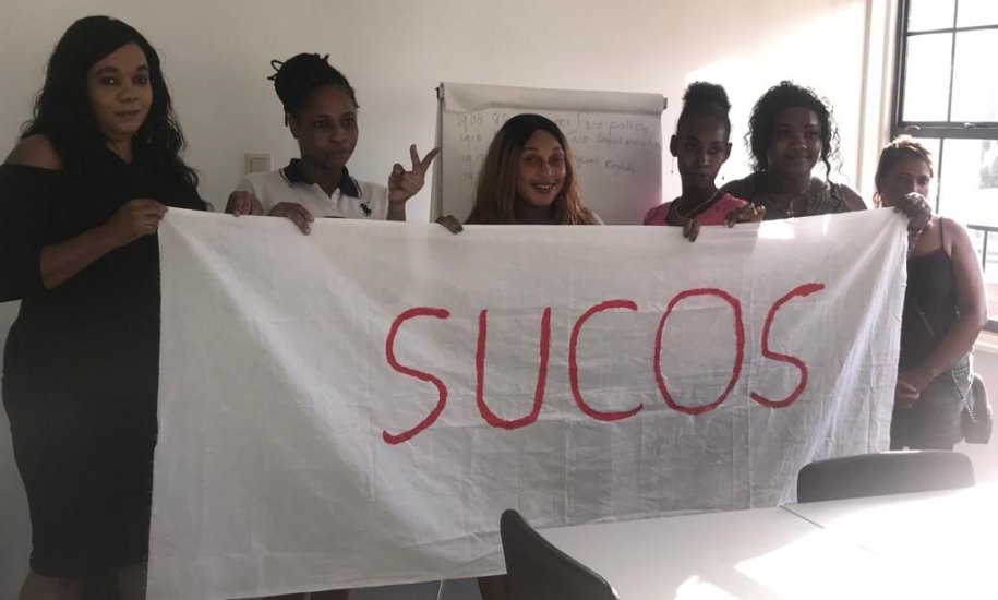 six women of colour stand in front of a table, next to a window, holding a large white banner that reads SUCOS in red writing.
