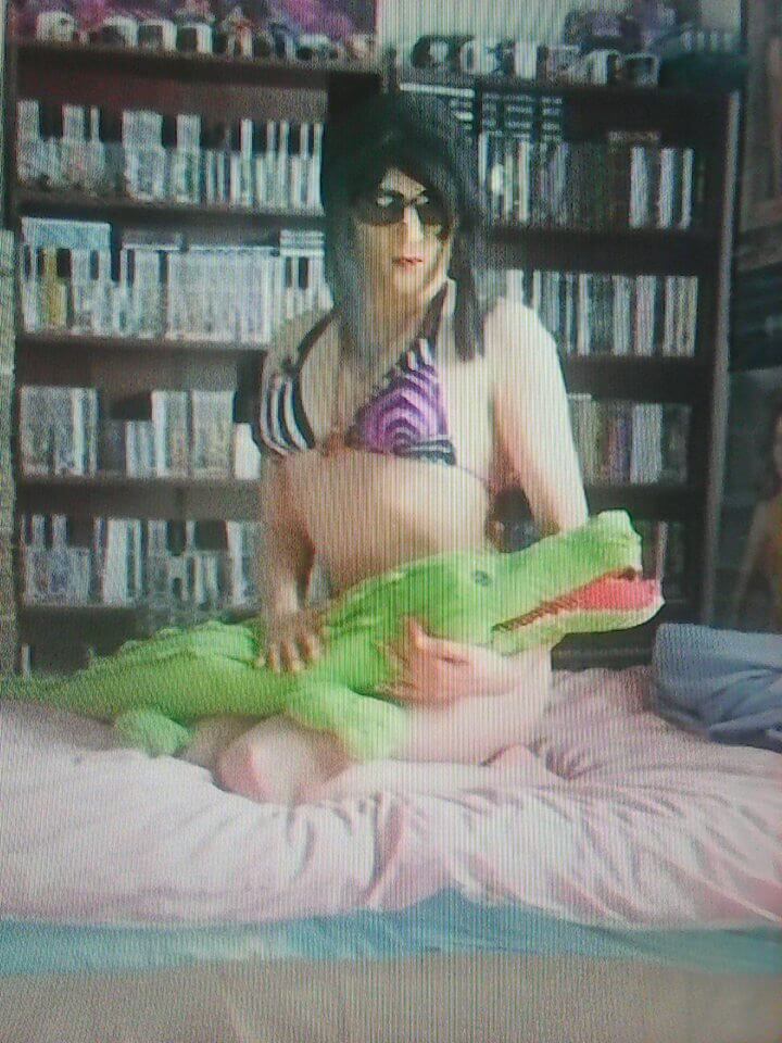 A person weaering a bikini top and sunglasses stare into the camera. They are holding a stuffed green crocodile across their lap as they kneel on a bed.