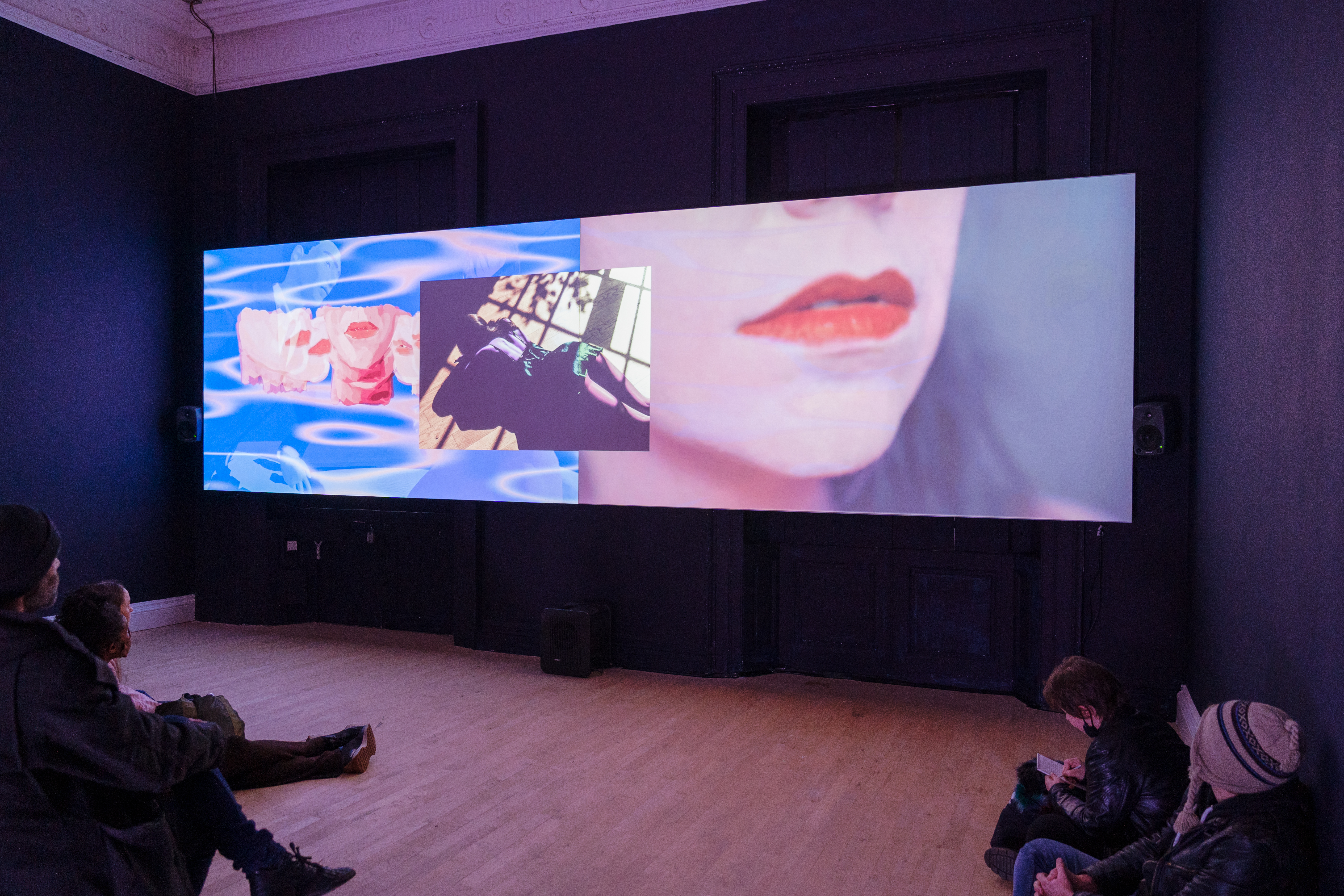 A darkened room showing a video projection of three images featuring the bottom of a face including red lips. People are sat around the edge of the room watching.