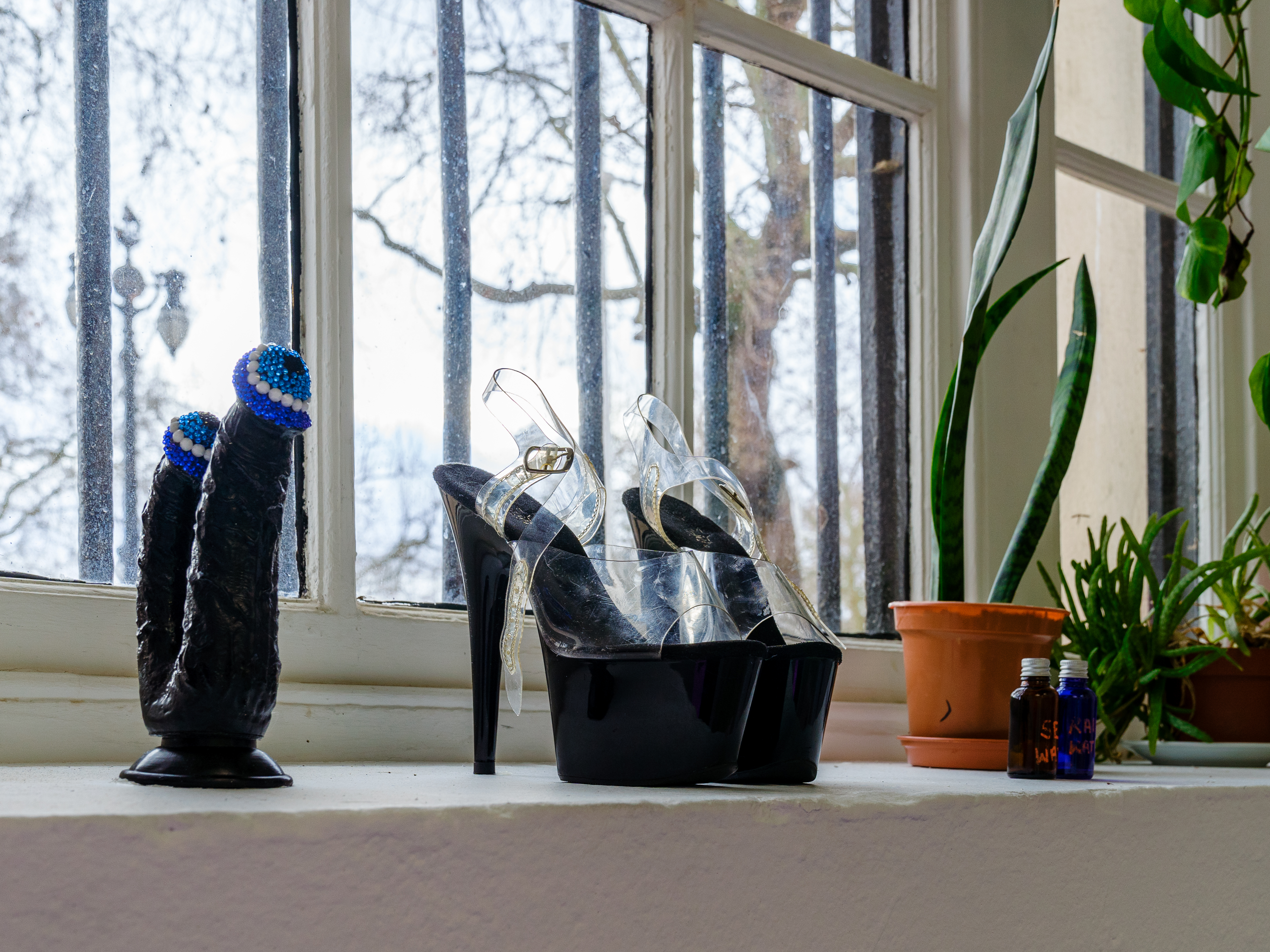 A window sill featuring a double ended dildo with crocheted hats, a pair of black pleaser heels and some plants.