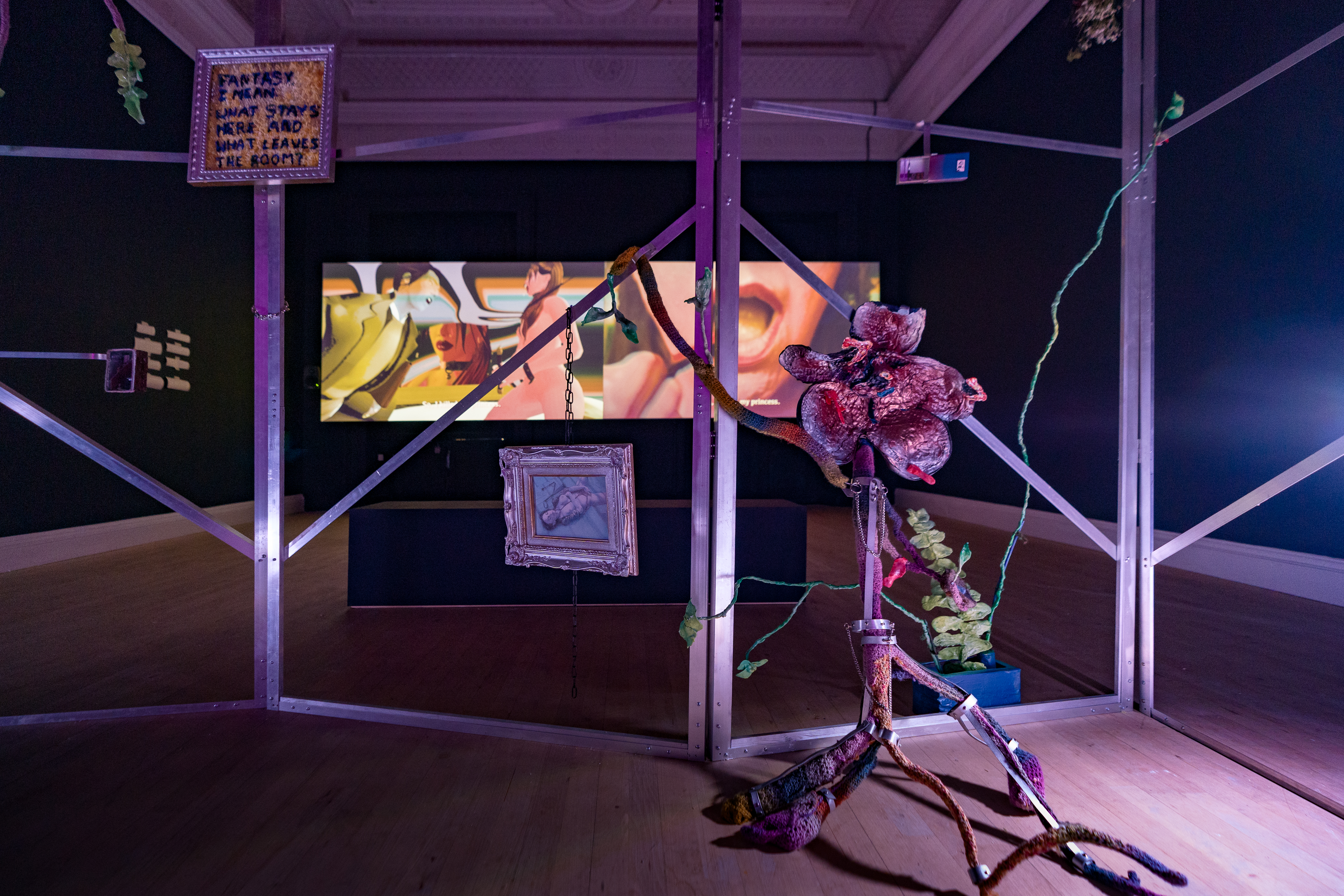 A gallery space featuring a video projection in the background, and a large metal frame in the foreground with picture frames attached to it and a fabric sculpture resembling a large flower.