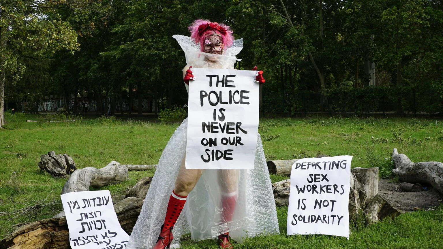 Artist Liad Hussein Kantorowicz poses in a field surrounded by trees and fallen branches. She wears a dress made of bubble wrap, red gloves and knee high socks, and a lace covering over her face. She holds up a large placard which says: 'the police is never on our side'.