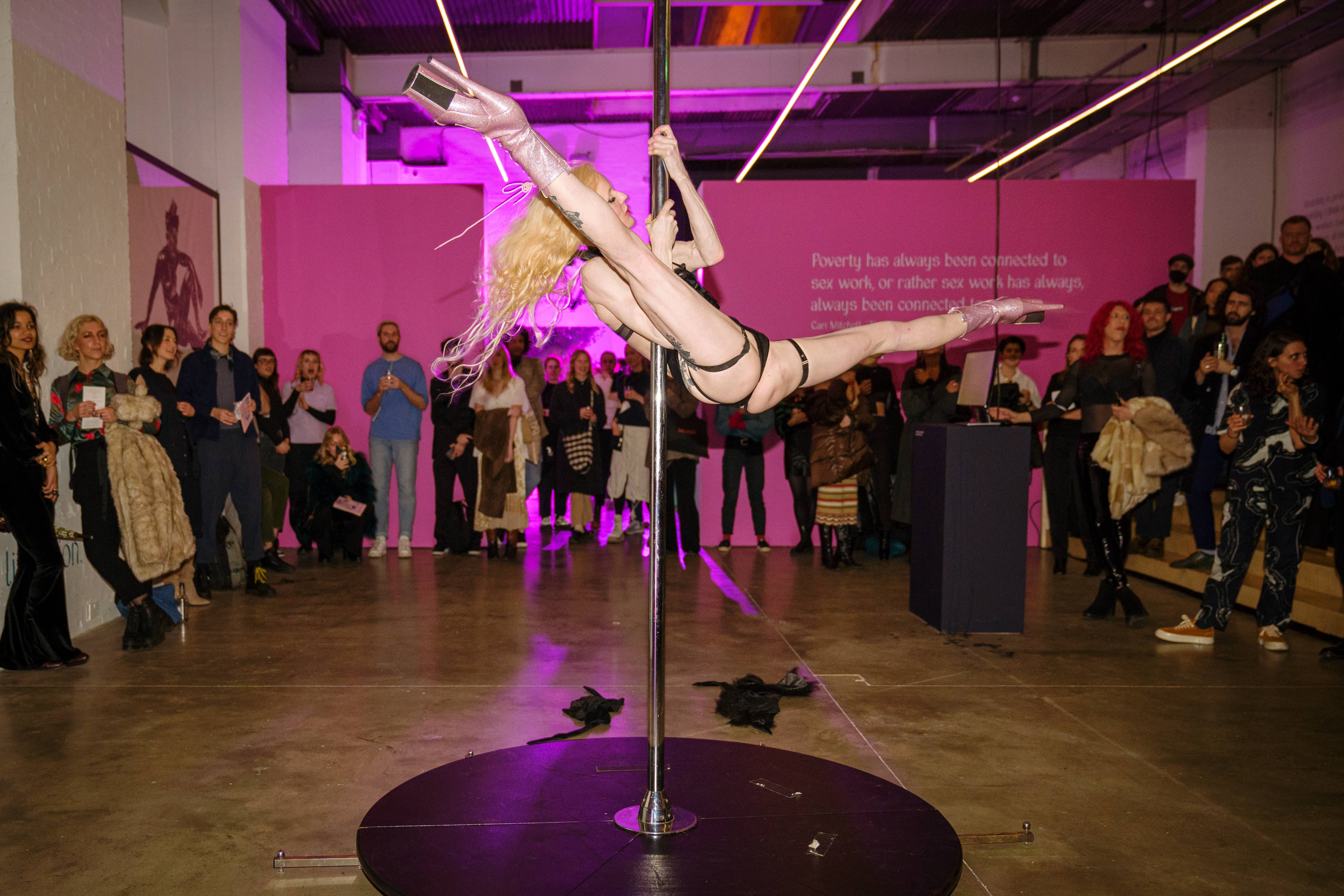 A crowd gathers in a gallery space to watch performer Tequila Rose. She is a thin, white person, holding herself high on a metal pole and has her legs open wide. She wears black underwear and pale pink high heeled boots.