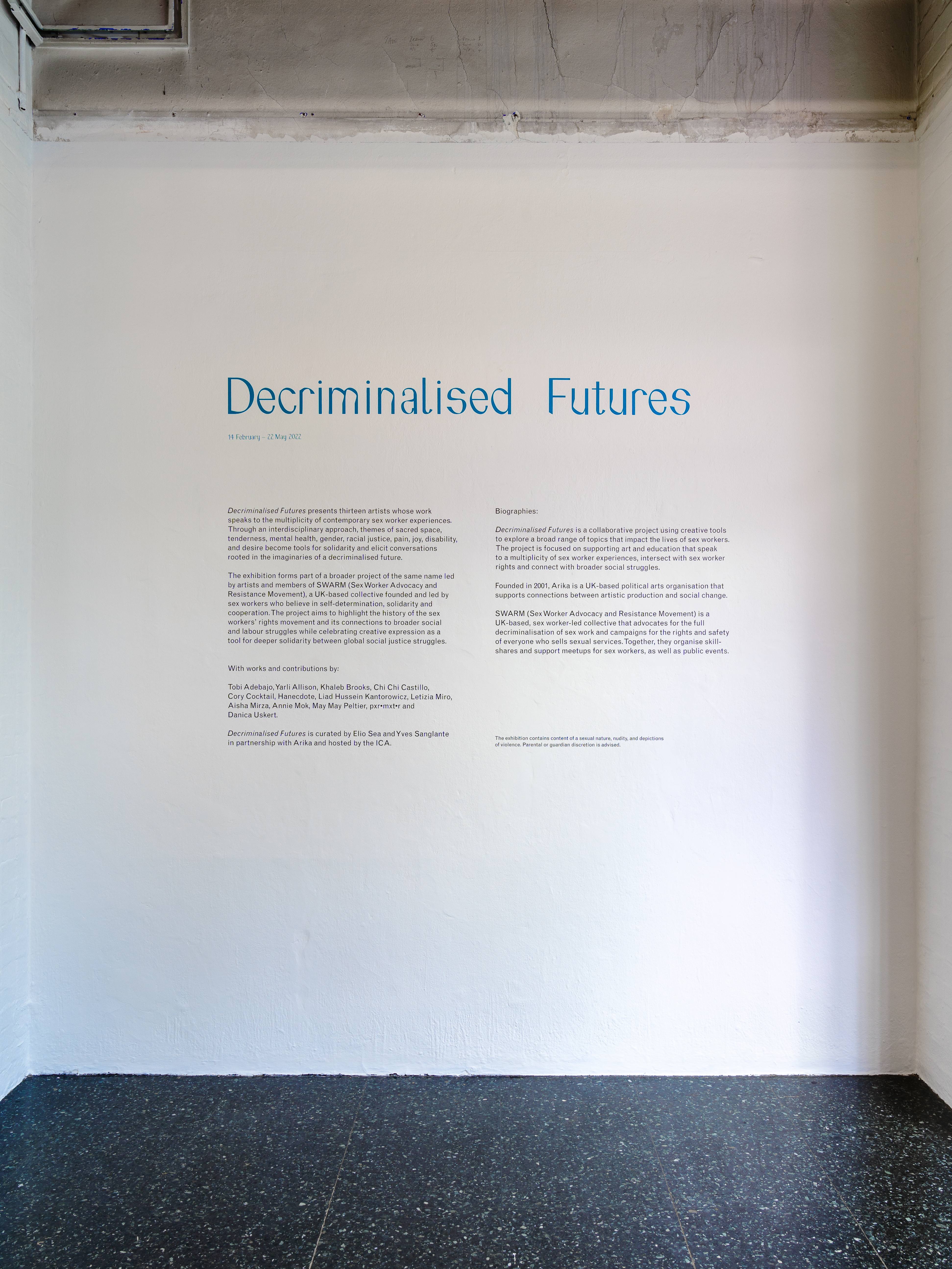 A white wall of a gallery space with a headline which reads 'Decriminalised Futures' in teal lettering. Underneath are several paragraphs of smaller, black text, describing the project and the exhibition.