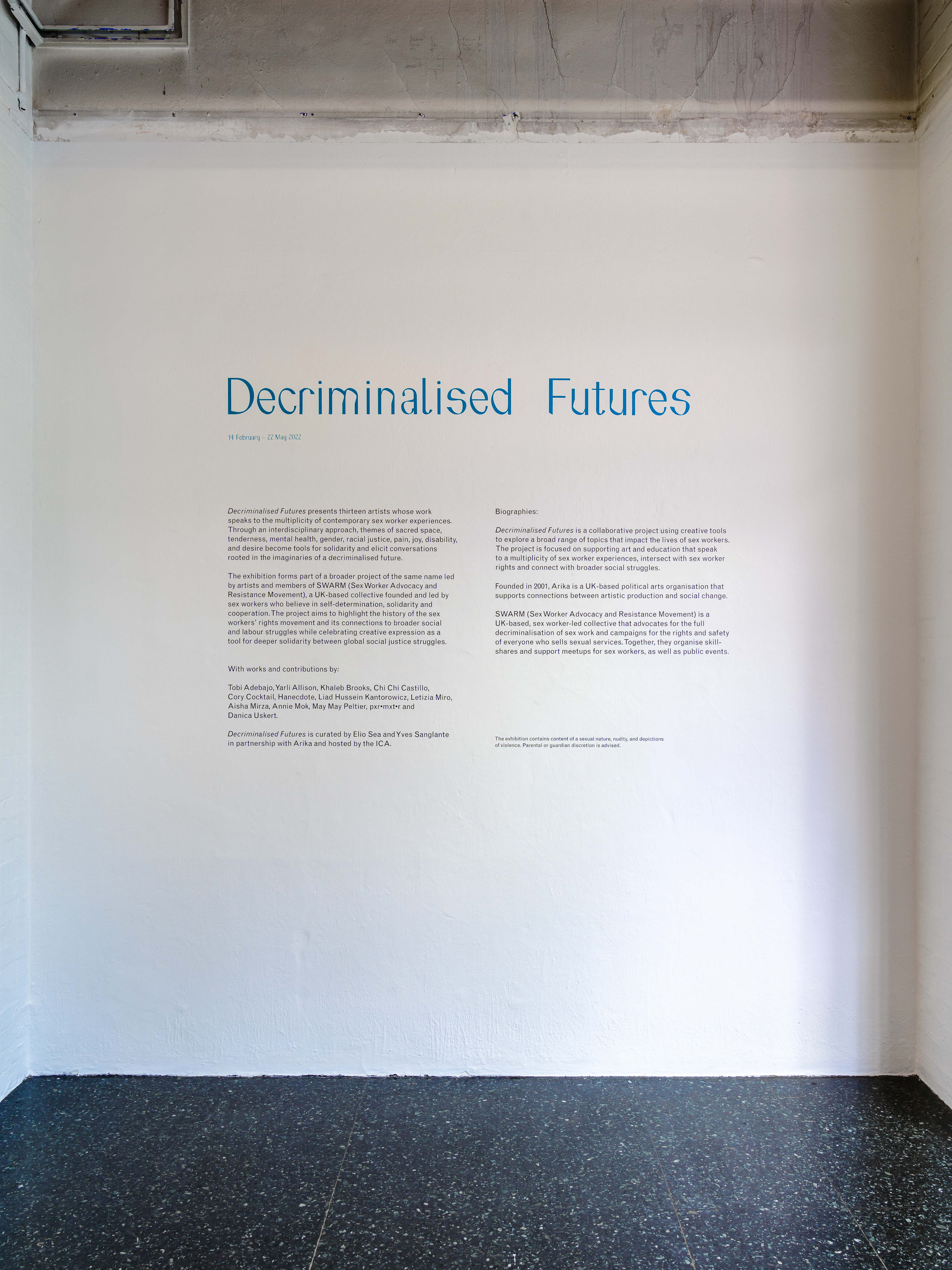 A white wall of a gallery space with a headline which reads 'Decriminalised Futures' in teal lettering. Underneath are several paragraphs of smaller, black text, describing the project and the exhibition.