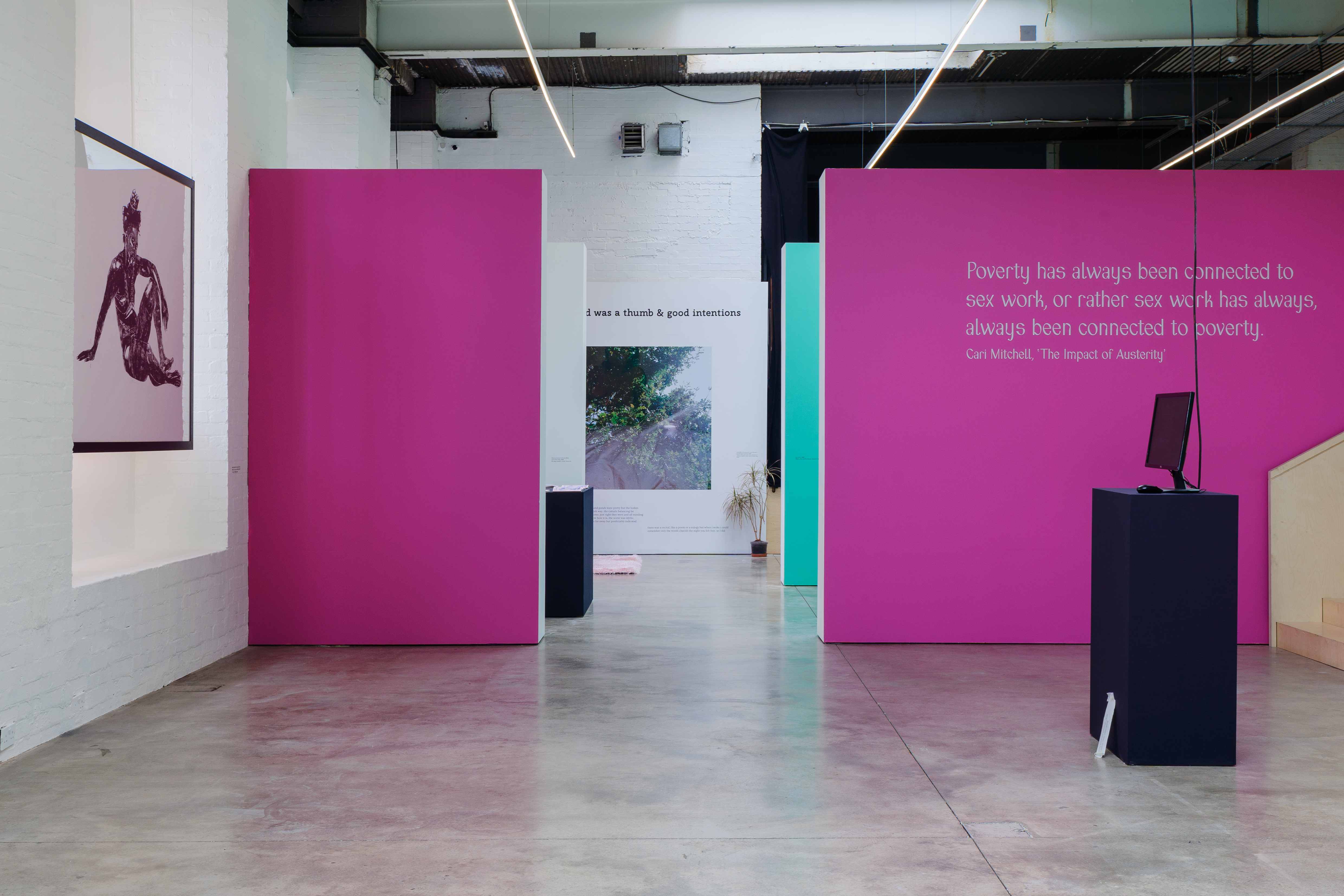 A gallery space is separated into three, with the image looking straight through them. A large figurative print is on the left wall, and a black plinth with a computer screen on the right side. The front facing wall is painted pink.