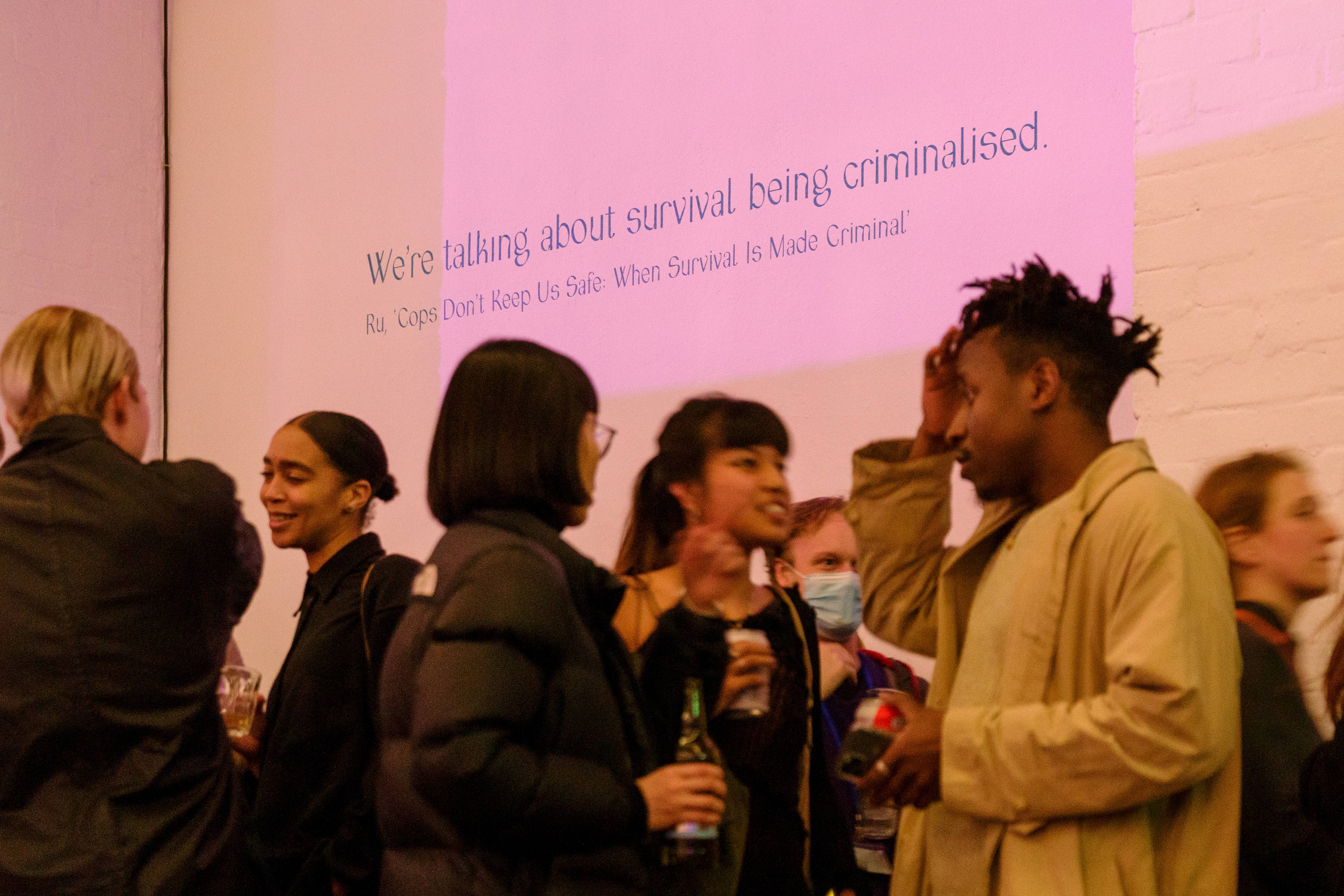 A group of exhibition attendees gather and talk underneath a large quote on the wall in teal lettering which reads: 'we're talking about survival being criminalised'.