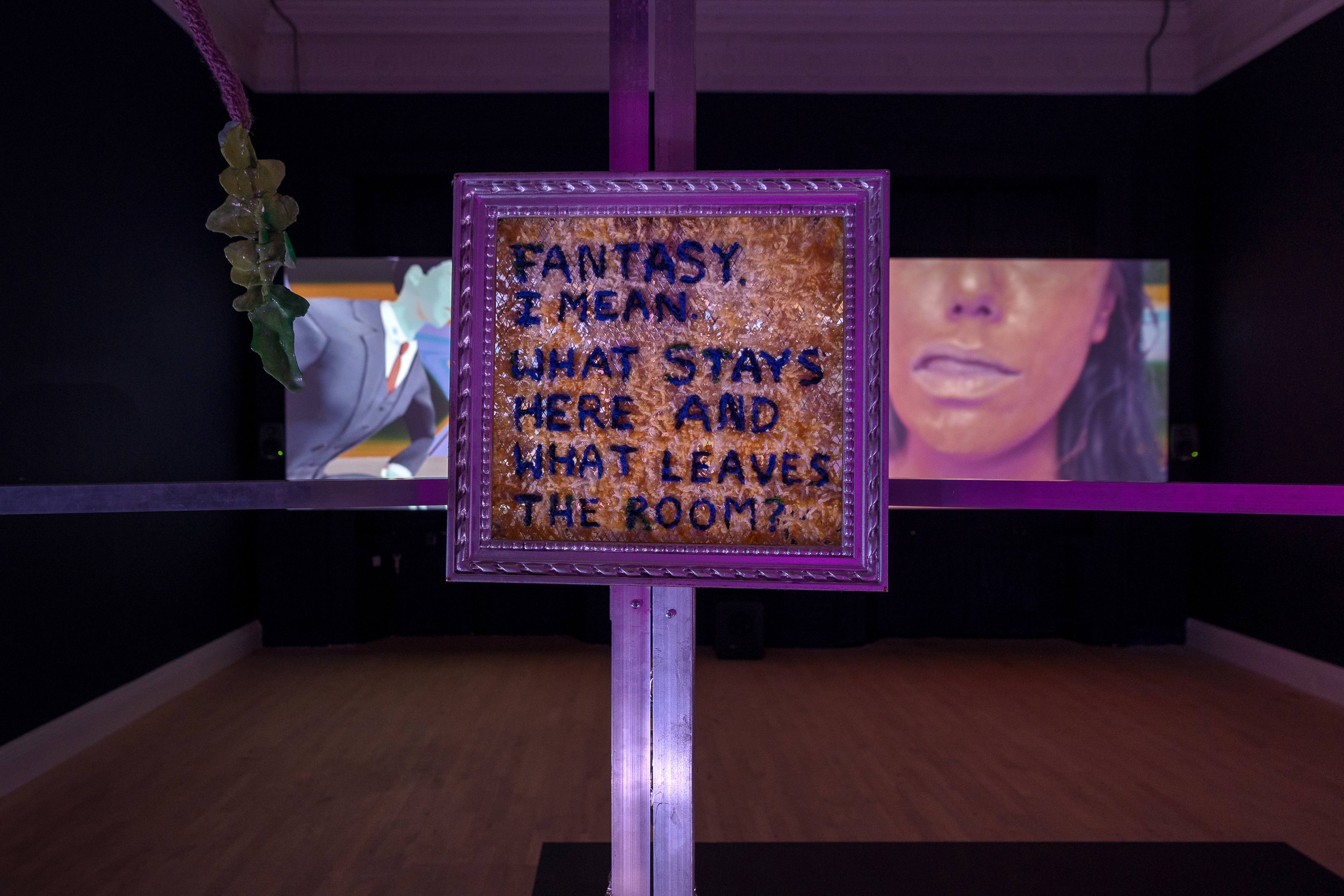 A dark room with a two screened video projection playing in the back wall. In the forefront is a framed text that says 'Fantasy I mean. What stays here and what leaves the room?' It is hung on metal frames.