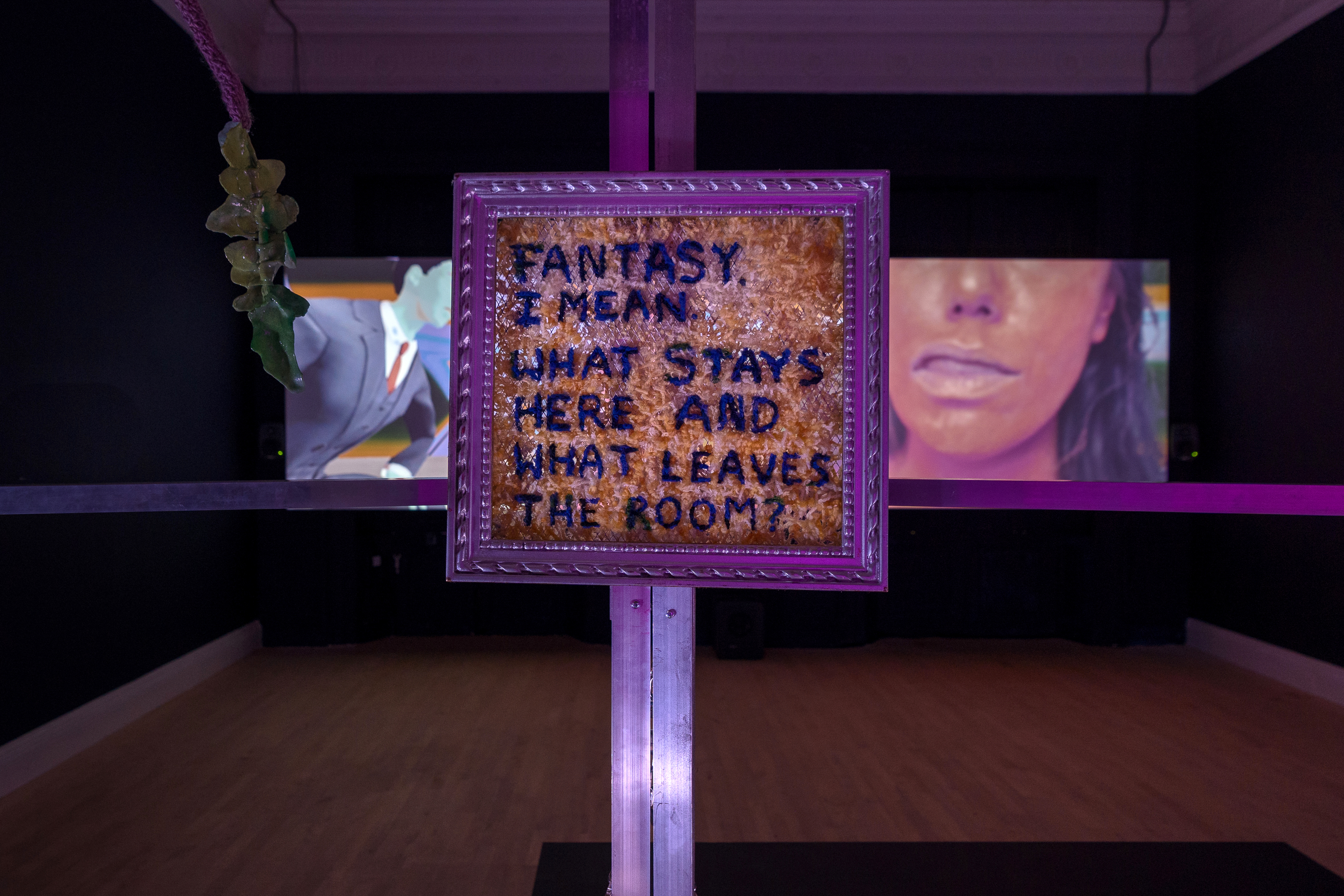 A dark room with a two screened video projection playing in the back wall. In the forefront is a framed text that says 'Fantasy I mean. What stays here and what leaves the room?' It is hung on metal frames.