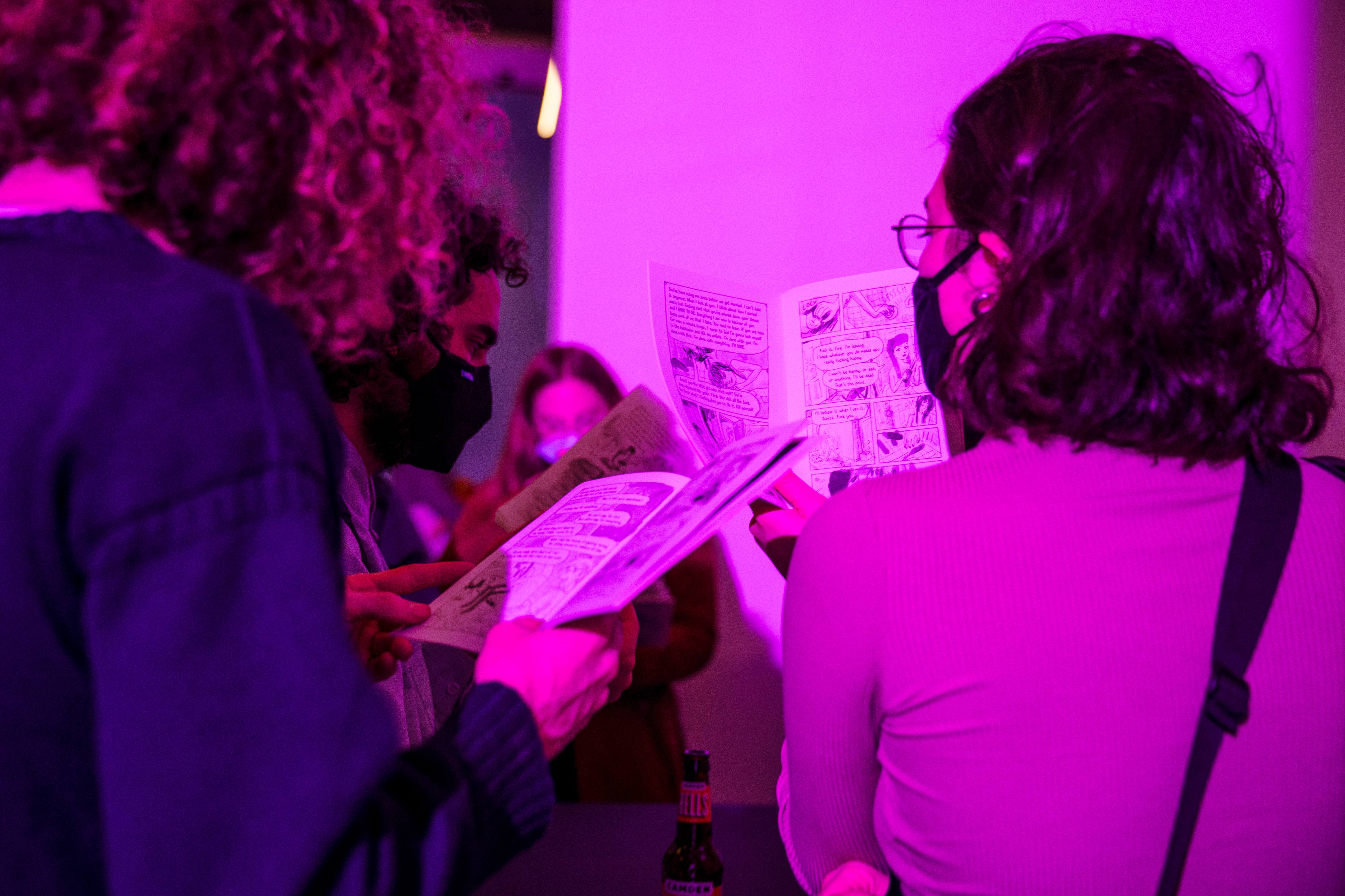 Attendees of the exhibition on opening night read through Danika Uskert and Annie Mok's comic. The attendees are wearing masks and standing in purple-pinkish light.