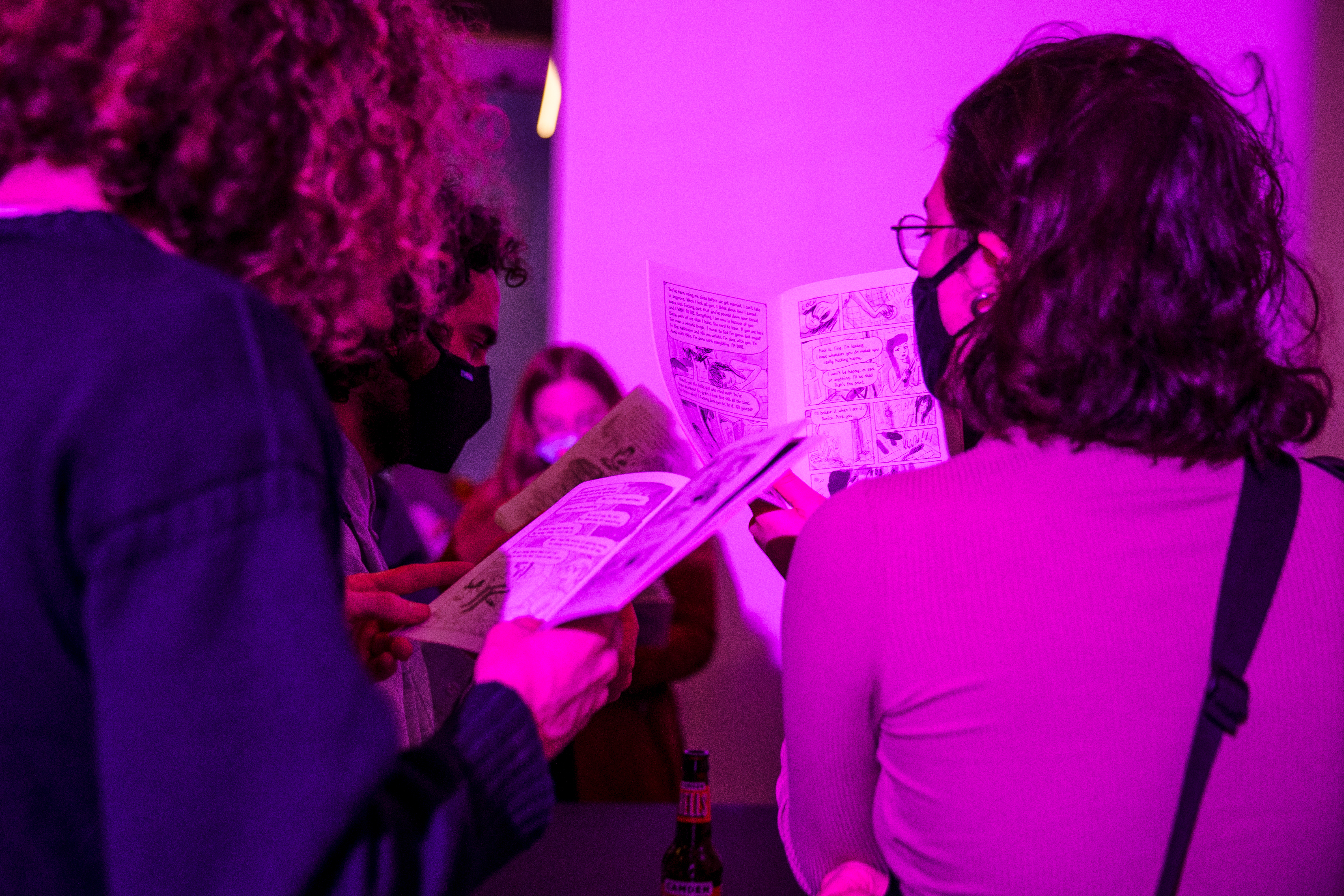 Attendees of the exhibition on opening night read through Danika Uskert and Annie Mok's comic. The attendees are wearing masks and standing in purple-pinkish light.
