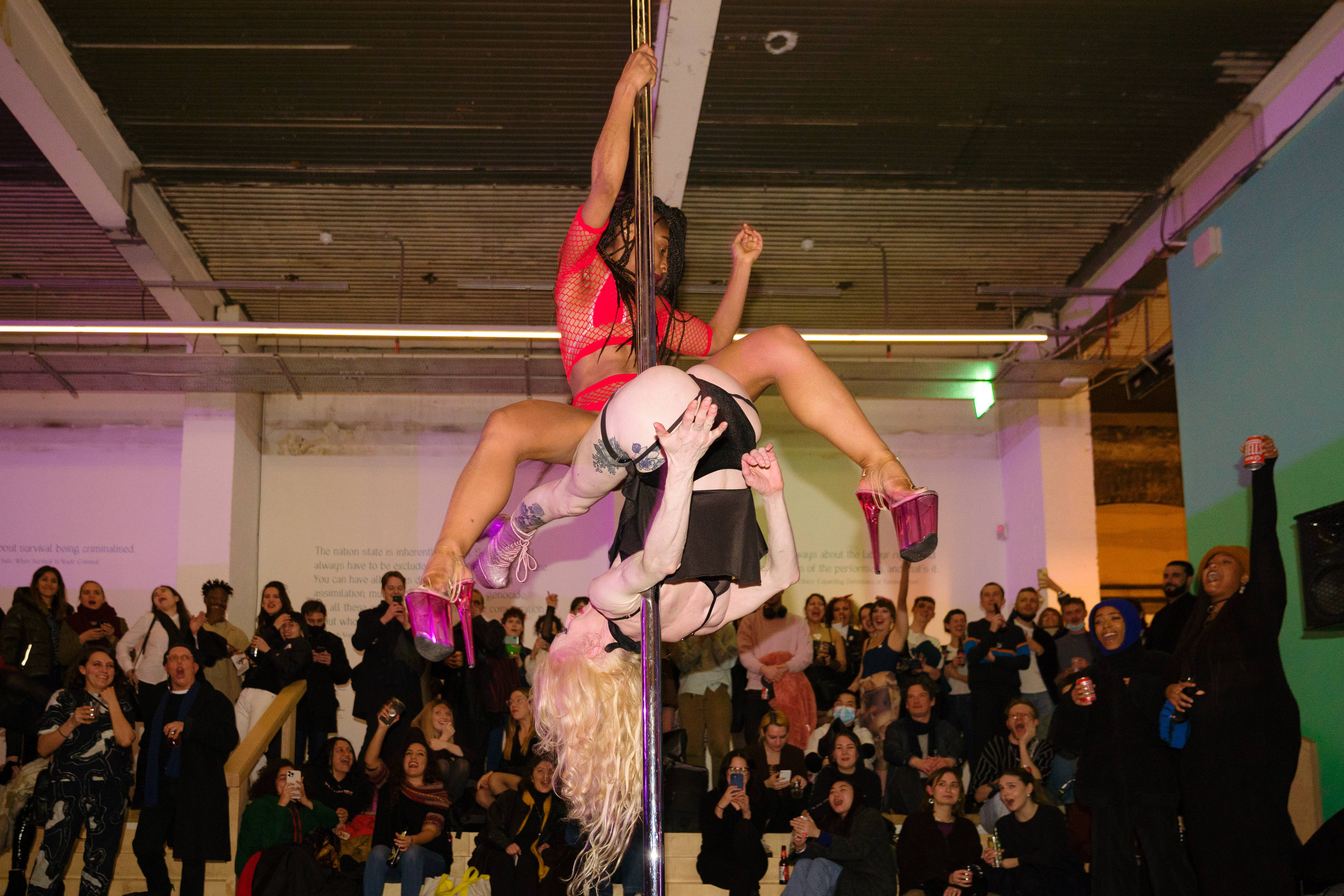 A large crowd cheer as they watch a pole performance by Tequila Rose and Sasha Diamond. Tequila is a white person in black underwear, and Sasha is a black person in a red fishnet top. They are both high up on the pole, Tequila is upside down and Sasha is holding herself up above her.