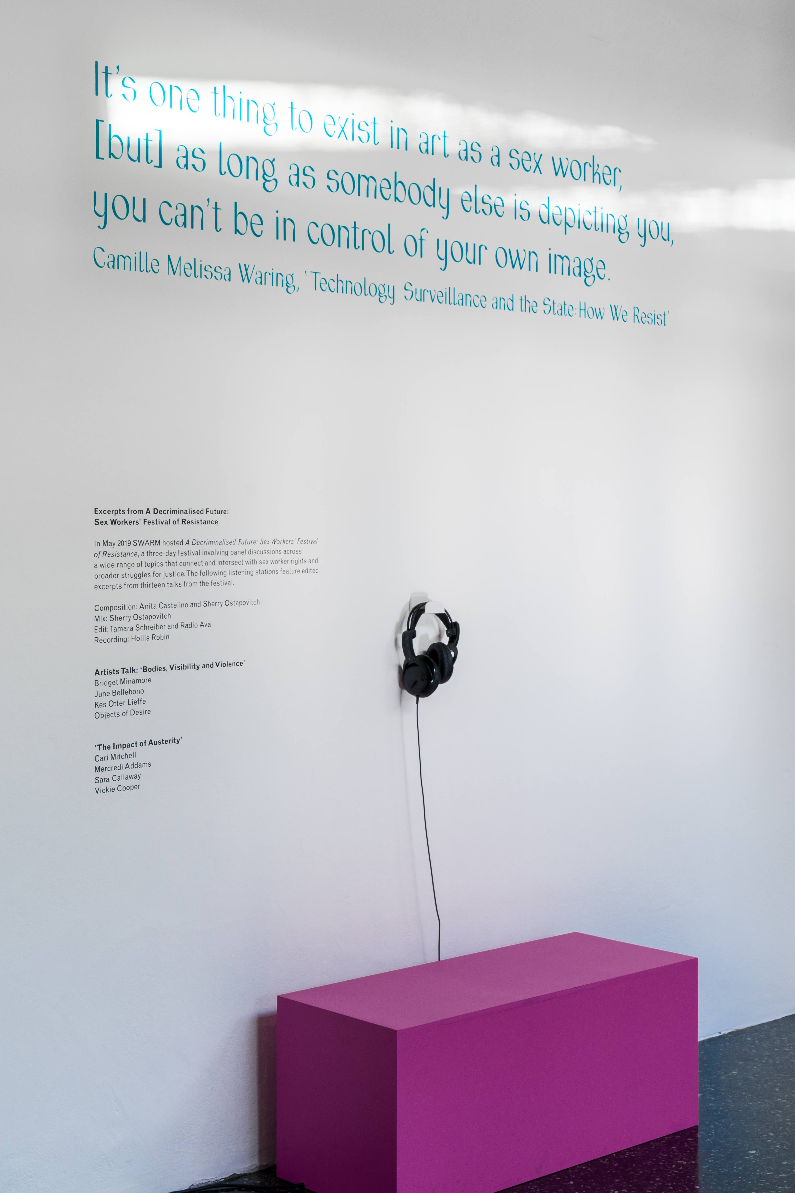 A white wall with a pink bench, and a pair of black headphones hanging from a hook. The text on the wall is a quote by Camille Melissa Waring which reads: 'it's one thing to exist in art as a sex worker, but as long as somebody else is depicting you, you can't be in control of your own image'.