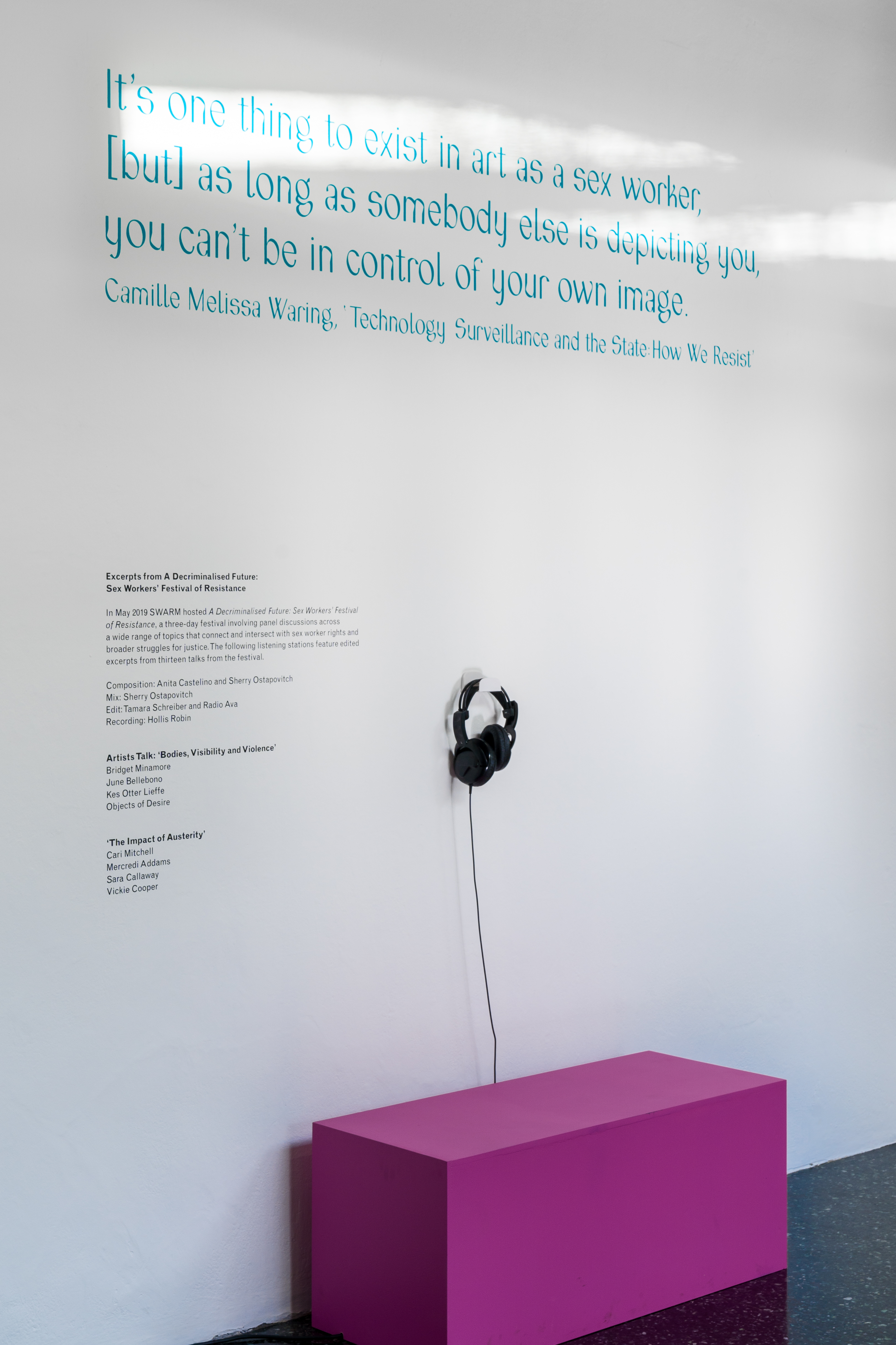 A white wall with a pink bench, and a pair of black headphones hanging from a hook. The text on the wall is a quote by Camille Melissa Waring which reads: 'it's one thing to exist in art as a sex worker, but as long as somebody else is depicting you, you can't be in control of your own image'.