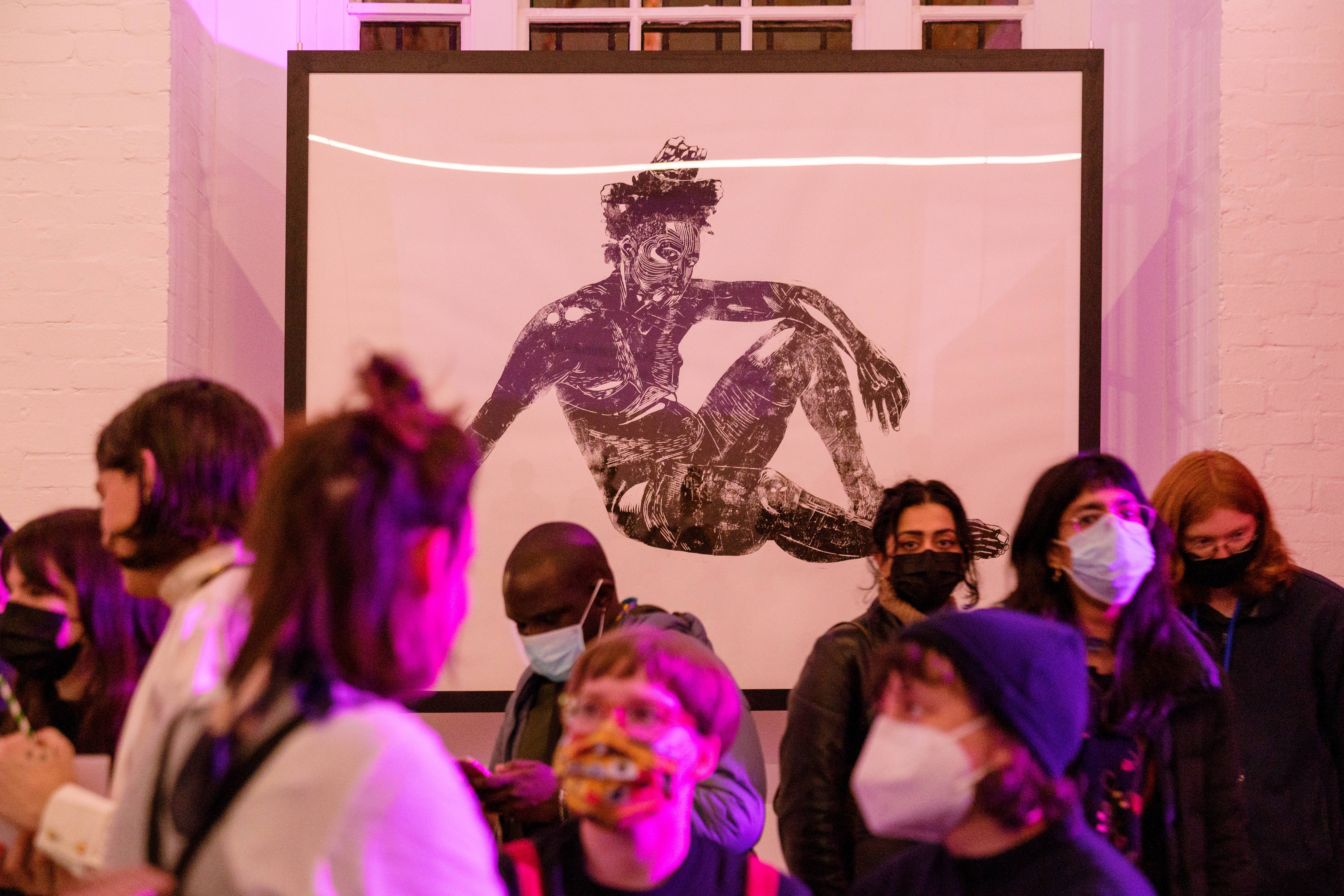 A crowd of masked-attendees at the opening night of the exhibition. On the wall above them is a large, framed, black and white lino print by Khaleb Brooks.