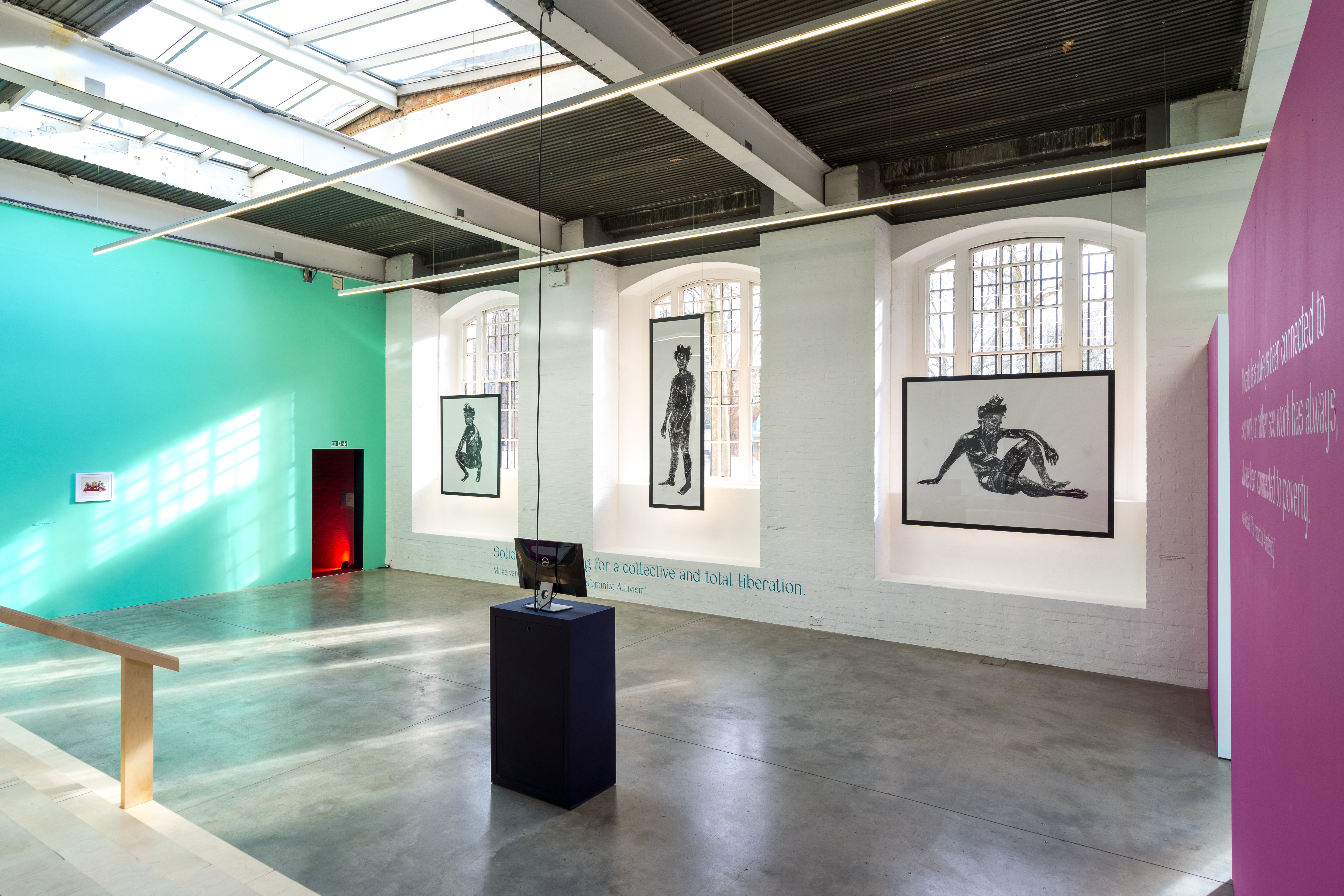 A large, bright gallery space shows three figurative prints by Khaleb Brooks, between three large arched gallery windows. A plinth is in the middle of the space, with a computer screen on it. The furtherest wall on the left is painted teal blue, and the one closest on the right is pink.