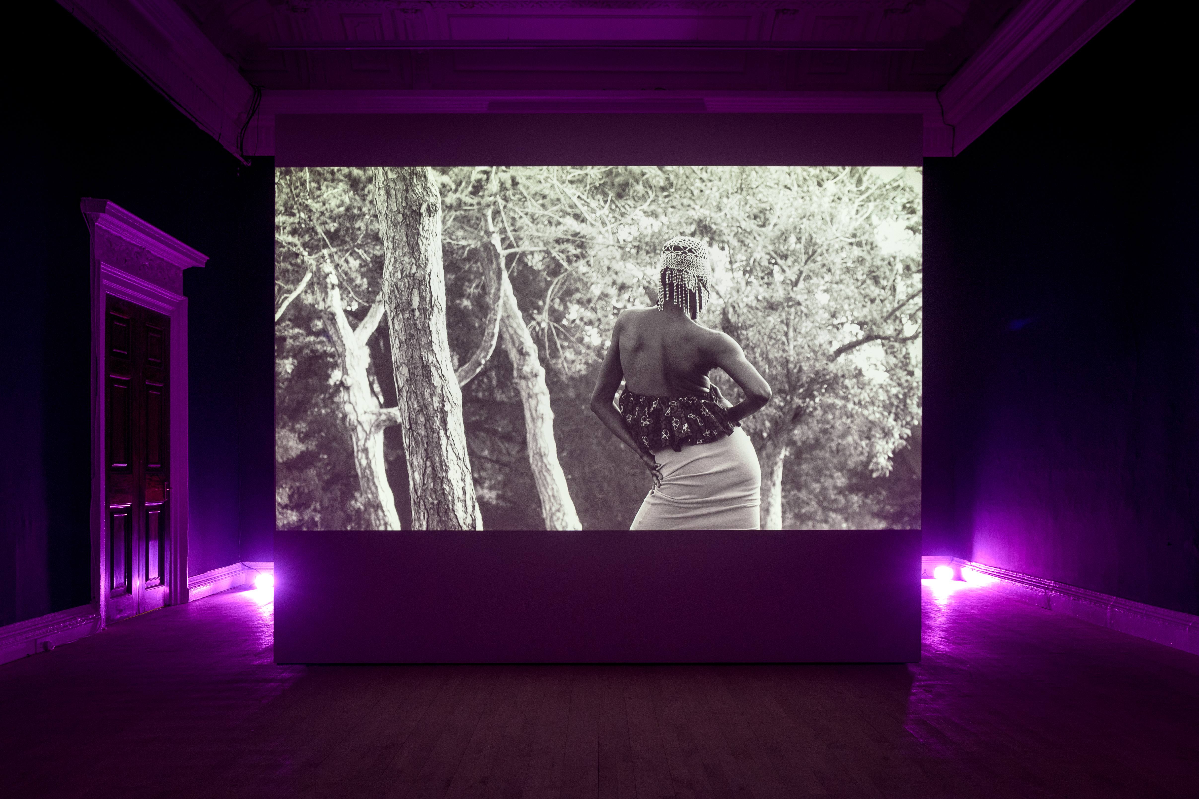 A darkened room with dim purple lights show a large projection of a black and white video, featuring the back of a person of colour who is standing amongst trees, wearing a headpeice and white skirt.