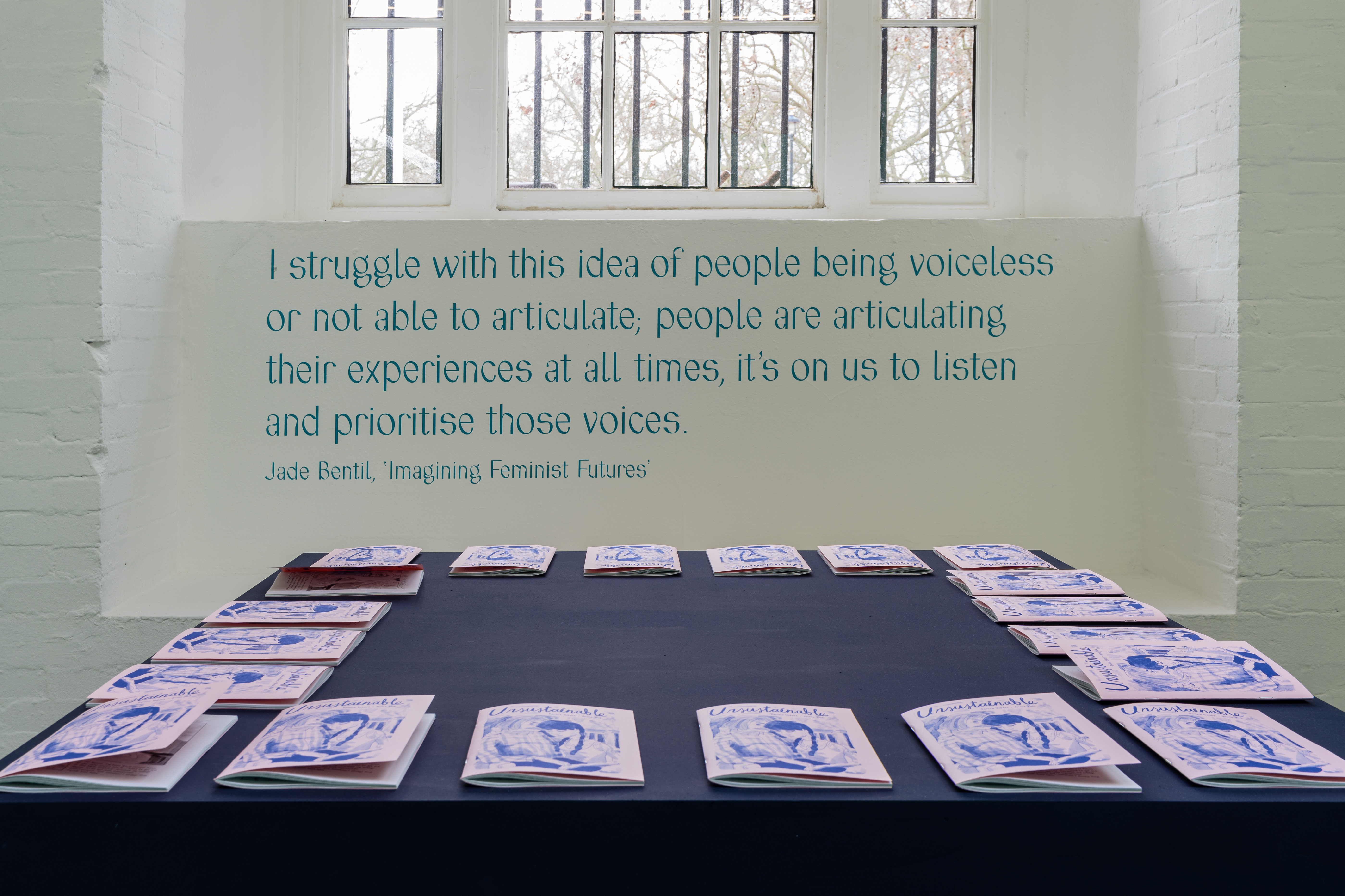 Zines are arranged on the edge of a square table covered in a dark tablecloth, in front of a window. Underneath the window text is printed 