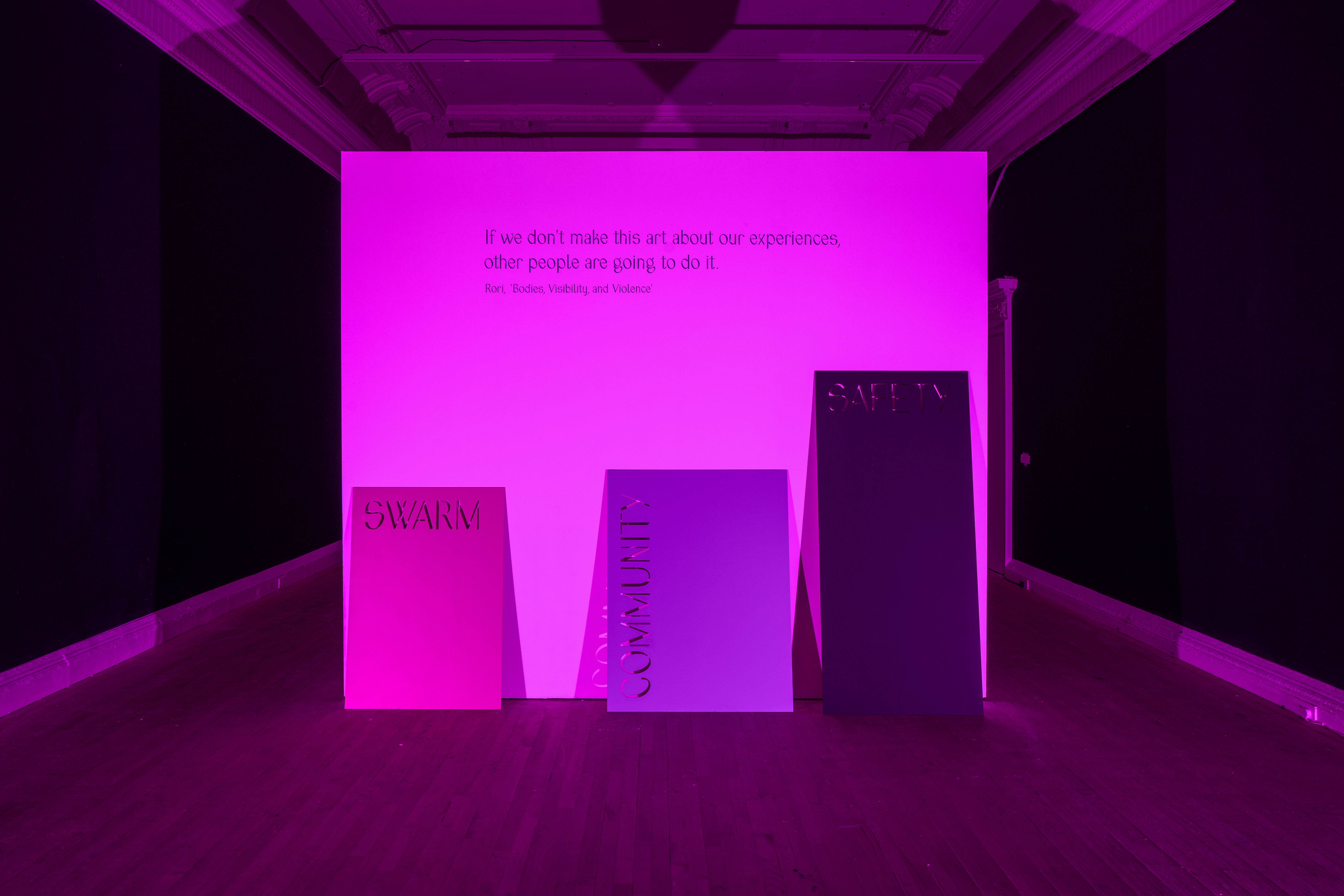 A darkened gallery room with dim purple-pink light. Three rectangular boards lean against one large white board, each saying 'SWARM', 'COMMUNITY' and 'SAFETY'. The large white board says 'if we don't make art about our experiences, other people will do it - Rori, Bodies, Visibility, and Violence'.