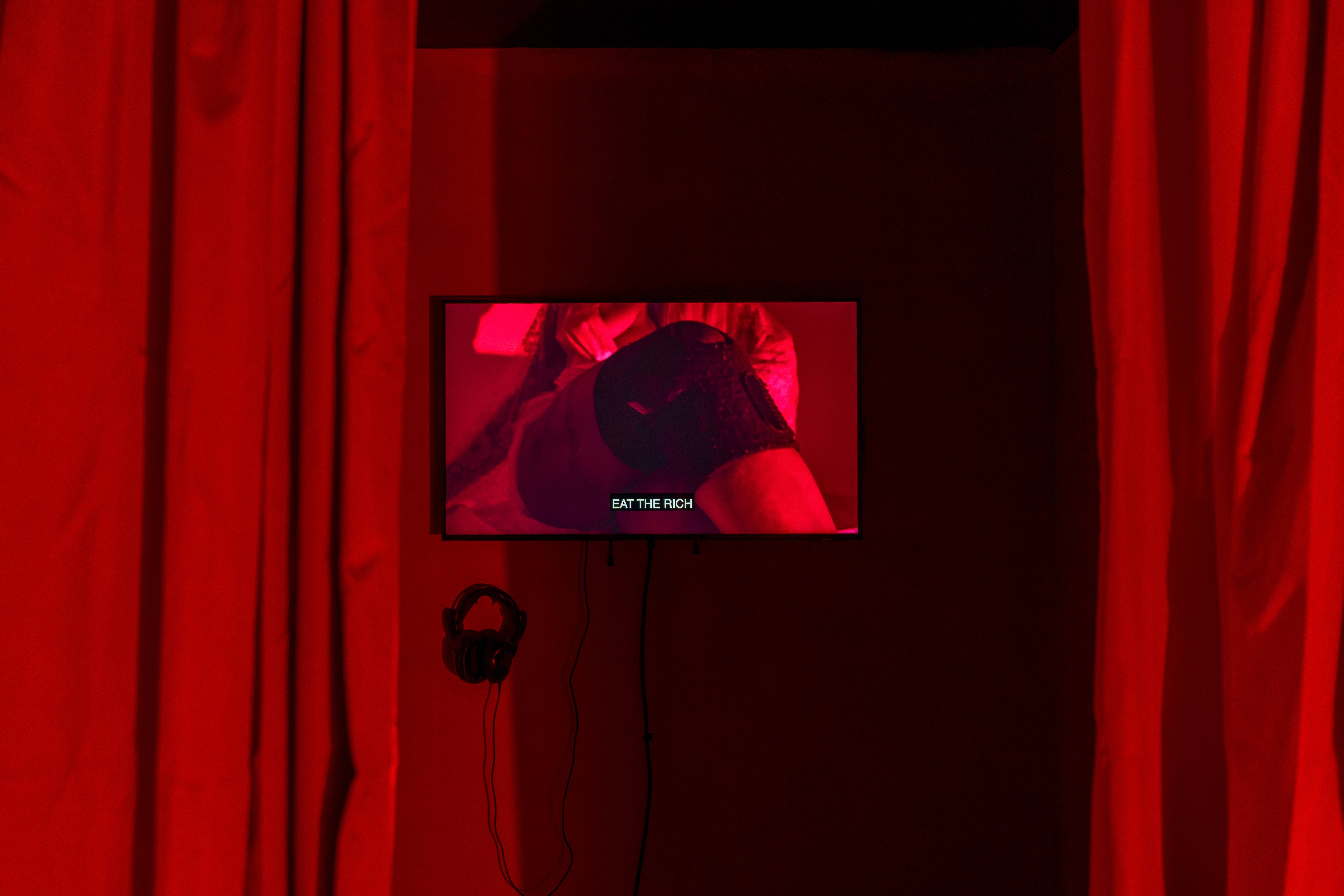 A close up show of a screen inbetween two red curtains. A red wash of light is prominent over the image, the image on the screen shows a knee in black kneepads, with the text 'eat the rich'.