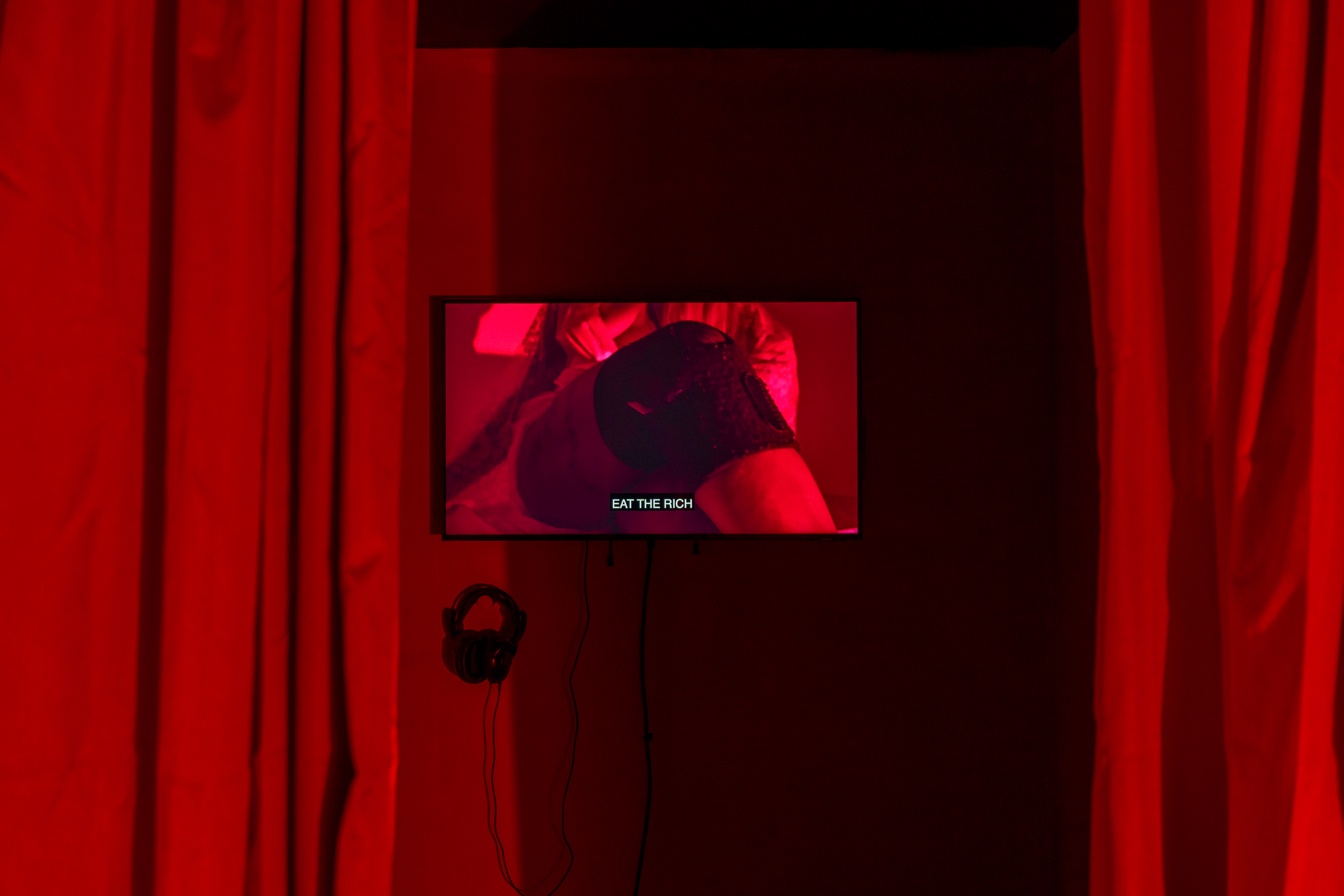 A close up show of a screen inbetween two red curtains. A red wash of light is prominent over the image, the image on the screen shows a knee in black kneepads, with the text 'eat the rich'.